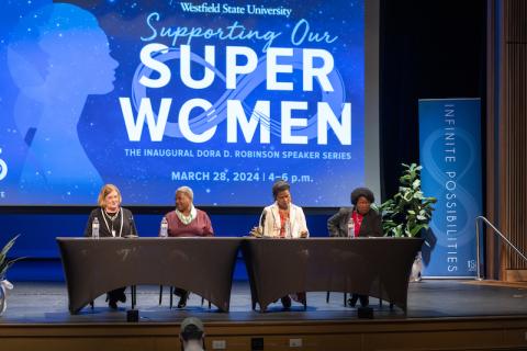 From the "Supporting Our Superwomen" symposium on March 28. Cheryl Woods Giscombe, Elizabeth Dineen, Kimberly Williams, and Phyllis Sharp sit on stage behind two black-clothed tables. Behind them is a blue graphic on a large projector, featuring "Supporting Our Superwomen" in blue and white letters. They look out at the audience during a Q&A session.