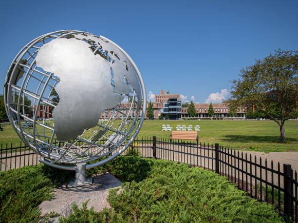 A stock photo of the silver campus globe. The sky above is blue, with green grass and vegetation around he globe itself, wrapped in by a black fence. Ely is in the distance, with a tree to the side of the frame.