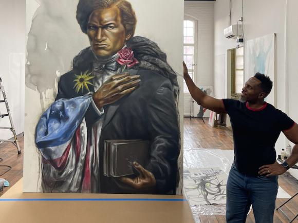 Dr. Imo Nse Imek, an Associate Art Professor at Westfield State. He is standing next to a painting of Frederick Douglass, and wears jeans and a black, short-sleeve shirt. The portrait is large and features Douglass holding a sunflower and rose in each hand. An American flag is draped around one of his arms. The background is Dr. Imeh's art studio and has white walls and a white, high ceiling.
