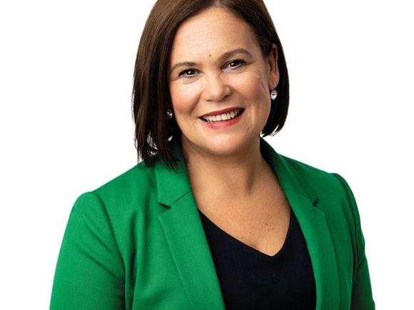Mary Louise McDonald, president of Ireland’s Sinn Fein political party. She is in a green, long-sleeved jacket with a black undershirt. Her hair is brown and cut to chin-length, and she smiles at the camera against a white backdrop.