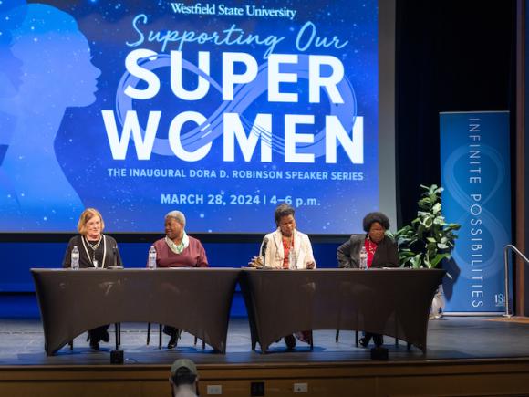 From the "Supporting Our Superwomen" symposium on March 28. Cheryl Woods Giscombe, Elizabeth Dineen, Kimberly Williams, and Phyllis Sharp sit on stage behind two black-clothed tables. Behind them is a blue graphic on a large projector, featuring "Supporting Our Superwomen" in blue and white letters. They look out at the audience during a Q&A session.