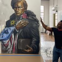 Dr. Imo Nse Imek, an Associate Art Professor at Westfield State. He is standing next to a painting of Frederick Douglass, and wears jeans and a black, short-sleeve shirt. The portrait is large and features Douglass holding a sunflower and rose in each hand. An American flag is draped around one of his arms. The background is Dr. Imeh's art studio and has white walls and a white, high ceiling.
